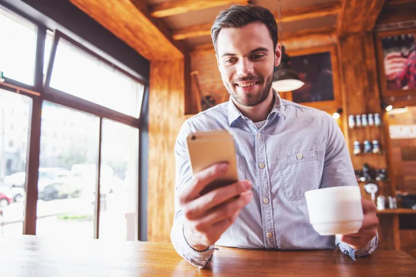 英俊的年轻商人是使用智能手机 捧着一杯咖啡和微笑 坐在酒吧柜台在现代城市的咖啡厅 — 图库照片
