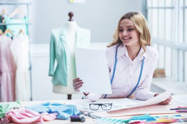 Güzel genç tasarımcı, bazı kroki inceleyerek ve dressmaking stüdyoda çalışırken gülümseyen, bir kağıt parçası tutuyor