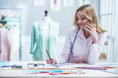 Güzel genç tasarımcı skeçler çiziyor, cep telefonu konuşuyor ve dressmaking stüdyosunda çalışırken gülümsüyor