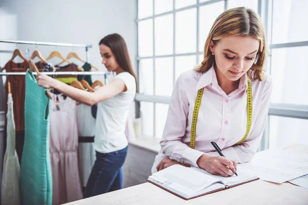 美丽年轻的设计师做笔记 而在缝纫工作室 在背景女孩工作选择礼服 — 图库照片