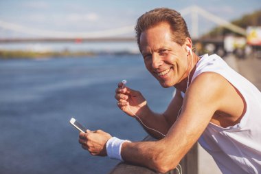 Yakışıklı orta yaşlı adam spor üniforma ve kulaklık smartphone kullanarak ve gülümseyerek kameraya bakarak müzik dinleme