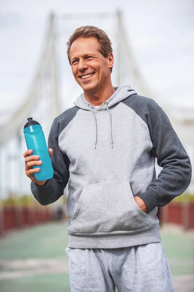 スポーツ フォームでハンサムな中年の男は ボトル入り飲料水と実行の朝に笑顔を保持しています — ストック写真