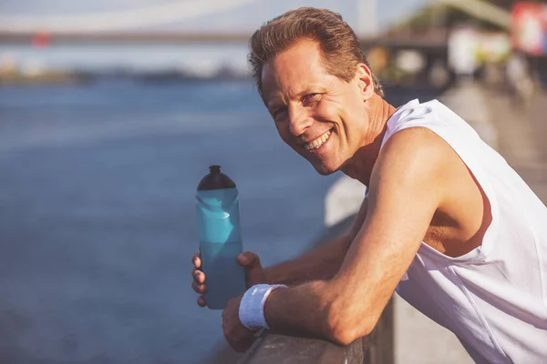 スポーツ フォームのハンサムな中年の男がカメラと笑みを浮かべて 朝のジョギング中に休憩を見て 水のボトルを保持してください — ストック写真