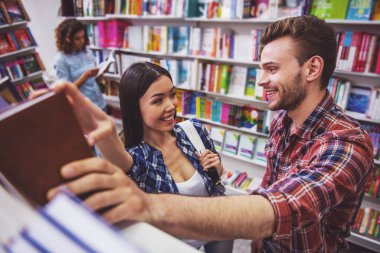 Yakışıklı adam ve güzel kız birbirlerine bakıyor ve kitapçıda aynı kitabı tutarken gülümsüyor, arka planda başka bir kız