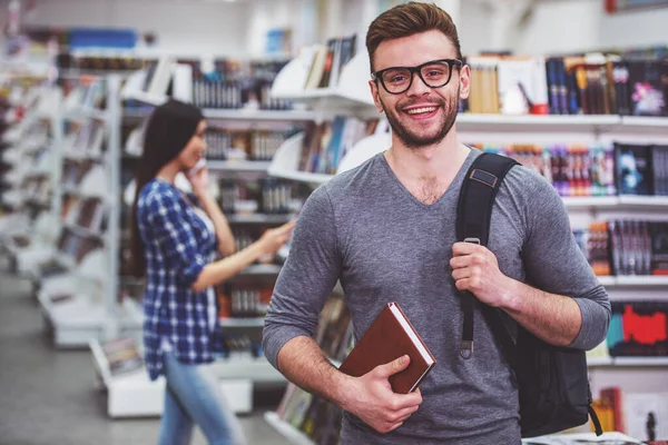 戴着眼镜的英俊学生拿着书 看着相机 微笑着站在书店里 在背景中的女孩正在选书 — 图库照片