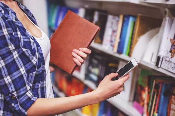 Przycięte Zdjęcie Atrakcyjna Dziewczyna Trzyma Książkę Pomocą Smartfona Stojąc Księgarni — Zdjęcie stockowe