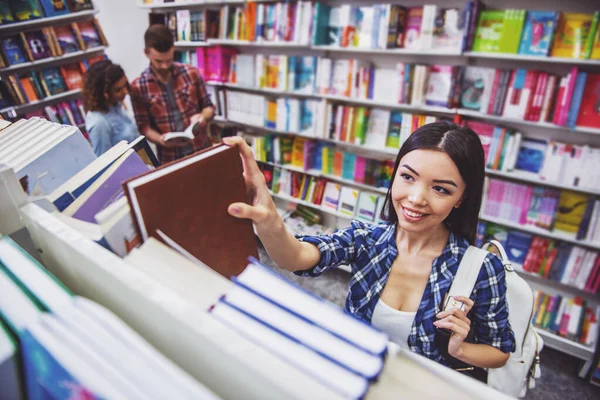 高角度看有吸引力的女孩微笑 而选择书在书店 年轻人的背景 — 图库照片