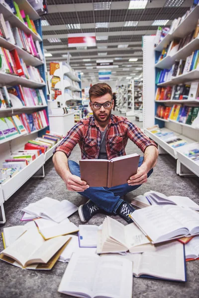 戴着眼镜的英俊惊讶的学生正在看书 而坐在地板上 在书店里放着大量打开的书 — 图库照片
