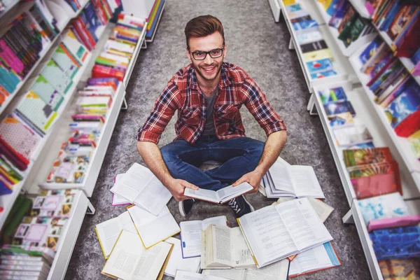 英俊的学生看书 看着相机 微笑着坐在书店打开的书的地板上的高角度视图 — 图库照片
