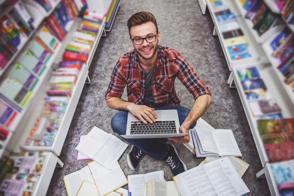 ノートパソコンを使ったハンサムな学生の高角図 書店で開かれた本の中で床に座りながらカメラを見て微笑む — ストック写真