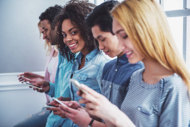 Akıllı telefonlar kullanarak ve üst üste otururken gülümseyen farklı milletlerden genç insanlar. Güzel Afro Amerikan kız kameraya bakıyor
