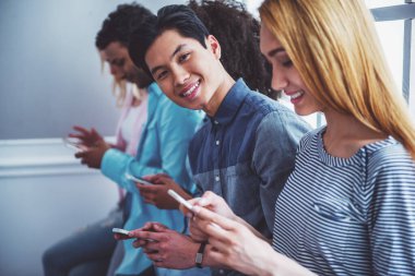 Akıllı telefonlar kullanarak ve üst üste otururken gülümseyen farklı milletlerden genç insanlar. Yakışıklı Asya'ya ait adam kameraya bakıyor
