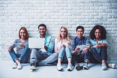 Güzel genç insanlar farklı milletlerden seyir kamera ve gülümseyerek, beyaz tuğla duvara oturmuş, becerikli, kullanmakta olduğunuz