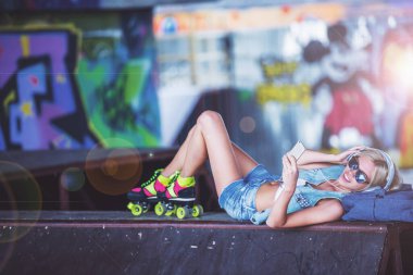 Güzel sarışın kız kulaklık ve bir smartphone kullanarak ve paten parkta yalan gülümseyen müzik dinlemek Silindirler yan görünüm