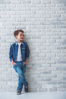 Kot pantolon giysiler içinde şık küçük çocuk tam uzunlukta portre uzağa bakıyor ve gülümseyerek, beyaz tuğla duvara karşı ceplerinde elleri ile ayakta