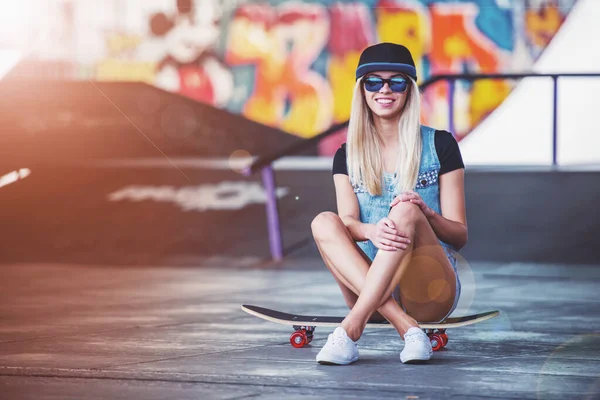 眼鏡とキャップで美しいブロンドの女の子はカメラを見て スケート公園で彼女のスケート ボードの上に座ってながら笑顔 — ストック写真