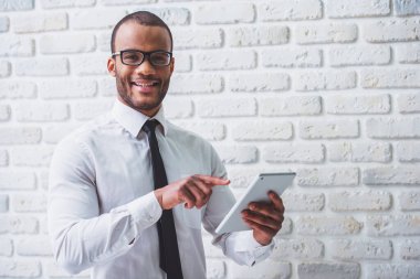 Klasik gömlek ve gözlük yakışıklı Afro Amerikalı işadamı tablet kullanıyor, kamera bakıyor ve gülümseyerek, beyaz tuğla duvara karşı