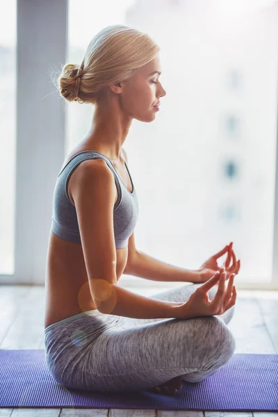 Side view of beautiful young woman in sportswear meditating while sitting in lotus pose on yoga mat against window