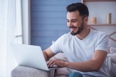 Yakışıklı bir dizüstü bilgisayar kullanarak ve evde kanepede otururken gülümseyen