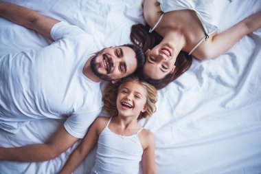 Güzel genç anne, baba ve onların kızı kameraya bakarak ve kafa kafaya yatakta yatıyordu süre gülümseyen Üstten Görünüm