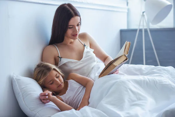 美しい若い母親は 自宅のベッドで寝ている小さな娘に本を読んでいます — ストック写真
