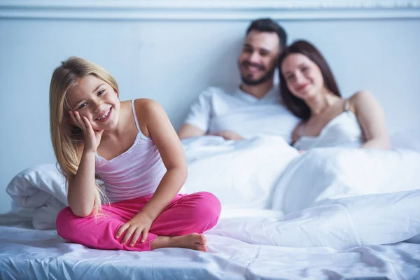 かわいい女の子のカメラ目線や笑顔のベッドの上に座っている間 バック グラウンドでの幸せな両親 — ストック写真