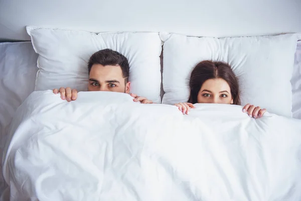 顶视图的美丽的年轻女子和她的男人躲在毛毯后面 看着相机躺在家里的床上 — 图库照片