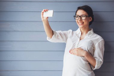 Gözlük güzel hamile iş kadını bir akıllı telefon kullanarak, bir yandan bir göbek üzerinde tutarak ve gülümseyen selfie yapıyor