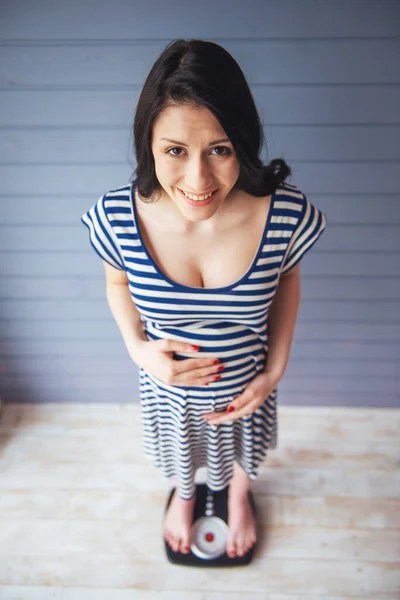 美しい妊娠中の女性を腹に手を維持するカメラを見て ニコニコと計量装置の上に立っての高角度を表示します — ストック写真