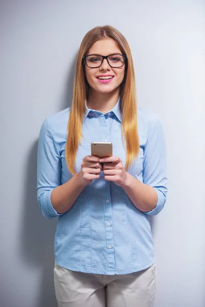 Attractive girl in classic clothes and in eyeglasses is using a smartphone, looking at camera and smiling, against gray background