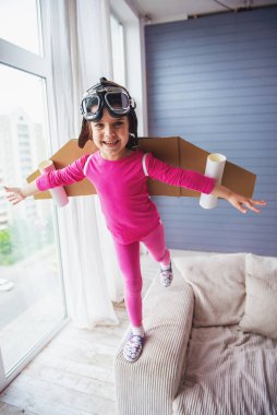 Sevimli küçük kız oyuncak kanatları ile bir pilot gibi giyinmiş gülümseyen ve evde oynarken kameraya bakıyor