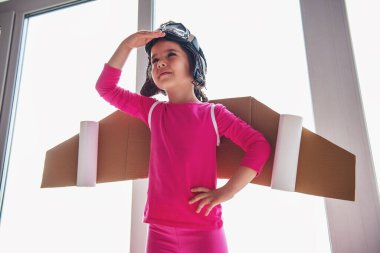 Oyuncak kanatlı bir pilot gibi giyinmiş sevimli küçük kız gülümsüyor ve pencerelere karşı duran, ufuk tarama