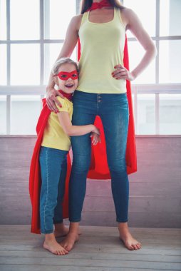 Güzel anne ve onun küçük kızı nın kırpılmış görüntü süper kahramanlar sarılma gibi giyinmiş, kamera bakarak ve gülümseyerek, pencerelere karşı ayakta