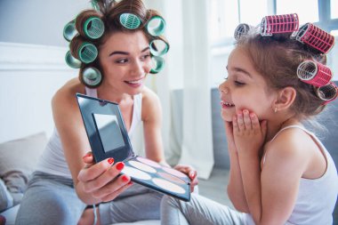 Güzel genç anne ve kızı saç curlers ile birlikte evde vakit. Anne kızı bir ayna gösterilen