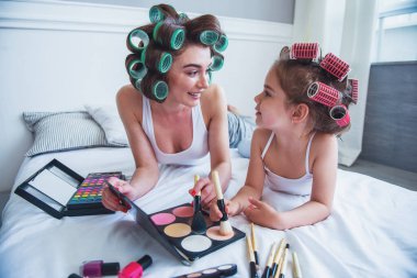 Güzel genç anne ve sevimli kızı ile saç curlers başları üzerinde makyaj, birbirine bakarak ve gülümseyen, yatağın üstüne oturuyor