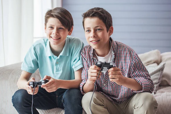 两个有吸引力的十几岁的男孩正在玩游戏控制台和微笑着坐在家里的沙发上 — 图库照片