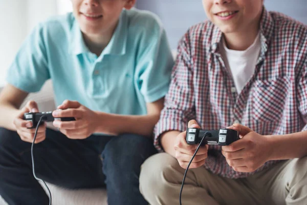 裁剪图像的两个有吸引力的十几岁的男孩玩游戏控制台和微笑着坐在家里的沙发上 — 图库照片