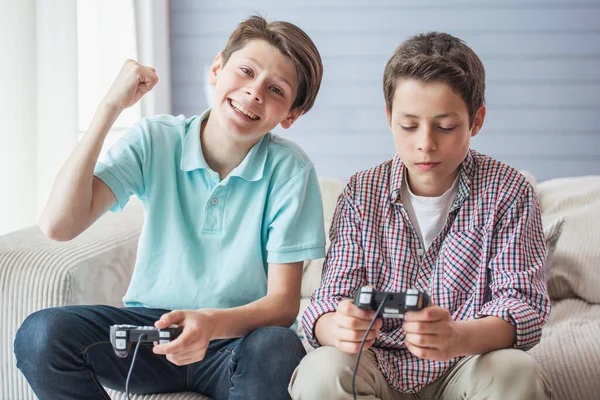 两个有吸引力的十几岁的男孩正在坐在家里的沙发上玩游戏控制台 一个家伙赢得 看着相机微笑 — 图库照片