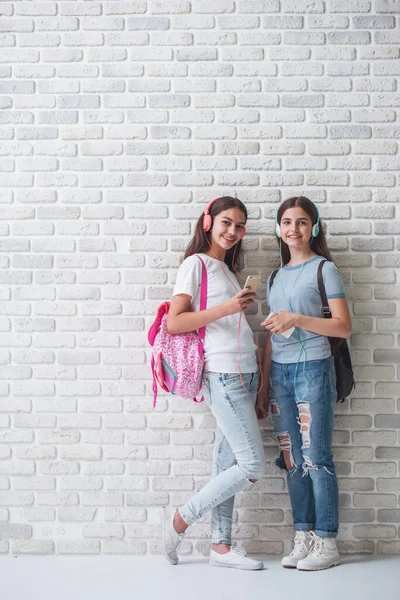 バックパックとヘッドフォンの 代の女の子は スマート フォンを使用して 笑って 白いレンガの壁に立って音楽を聴いています — ストック写真