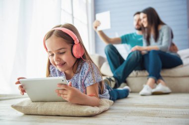 Kulaklık sevimli küçük kız bir tablet kullanıyor ve gülümseyerek, arka planda anne ve babası da bir tablet kullanıyor, evde kanepede oturuyor