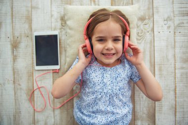 Kulaklıklı sevimli küçük kızın üstten görünümü tablet kullanarak müzik dinlemek, kameraya bakmak ve ahşap zeminde yatarken gülümseyen