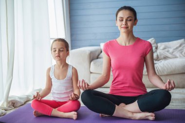 Güzel genç anne ve sevimli küçük kızı gülen ve lotus poz yoga mat evde otururken meditasyon
