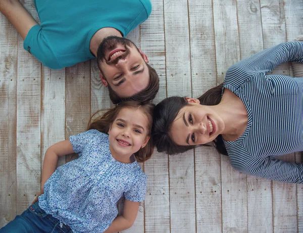 美丽的年轻父母和他们可爱的小女儿看着相机 微笑着 躺在木地板上 — 图库照片