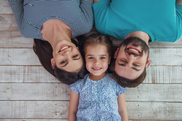美丽的年轻父母和他们可爱的小女儿看着相机 微笑着 躺在木地板上 — 图库照片