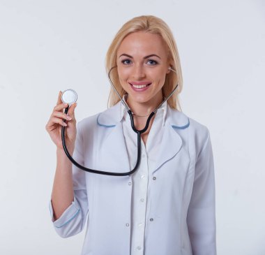 Kameraya bakarak ve beyaz arka plan üzerinde izole bir stetoskop tutarken gülümseyen beyaz paltolu güzel kadın doktor portresi