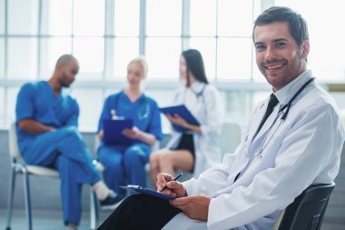 Yakışıklı doktor notlar, kameraya bakıyor ve gülümseyen, üç tıp doktorları bahsediyorsun arka planda