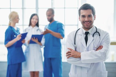 Yakışıklı doktor kameraya bakıyor ve gülümsüyor, çapraz kollarında üç doktor konuşuyor ve Notlar arka plan ayakta