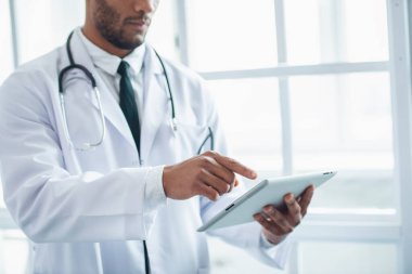 Yakışıklı kırpılmış görüntü Afro Amerikalı doktor ofisinde ayakta tablet, kullanma beyaz ceket