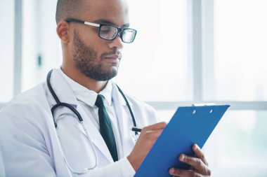Beyaz önlük ve gözlük yakışıklı Afro Amerikalı doktor ofisinde ayakta notlar, yapıyor