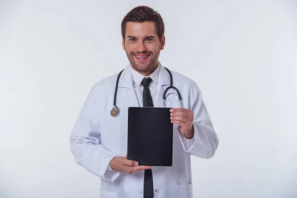 Przystojny Lekarz Białym Płaszczu Pokazuje Tablet Patrząc Kamery Uśmiechnięty Białym — Zdjęcie stockowe